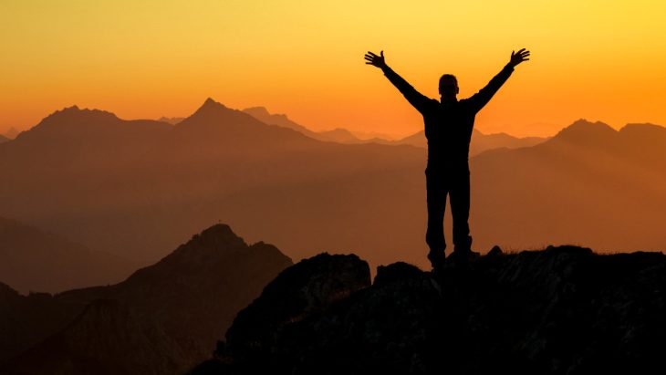 A person throws their hands up with excitement and achievement at the top of a mountain