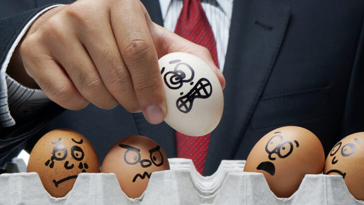 A man in a suit places an egg with an anxious face drawn onto it into a carton