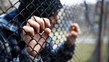 A sad-looking person in a hoodie grips a chain link fence