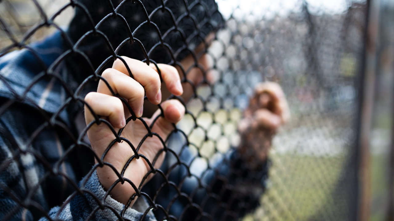 A sad-looking person in a hoodie grips a chain link fence