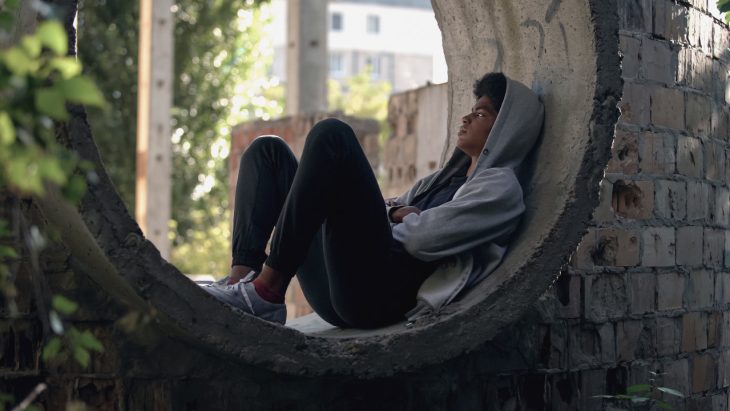 A depressed looking man in a hoodie sits in a hole in a brick wall
