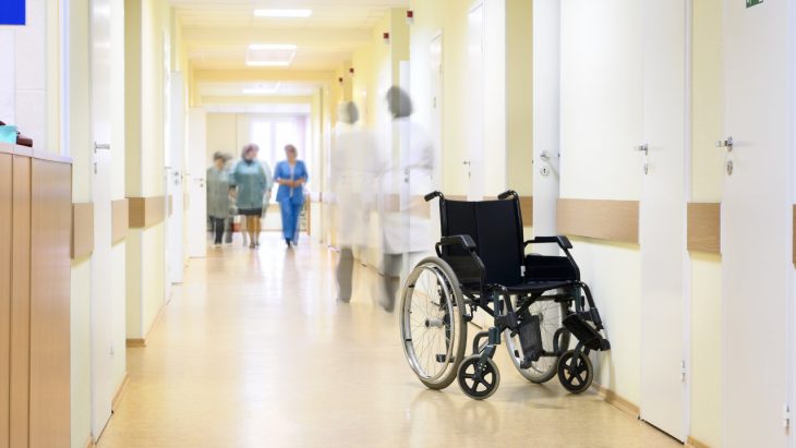 A view inside a hospital, with and empty wheelchair and care providers in the background