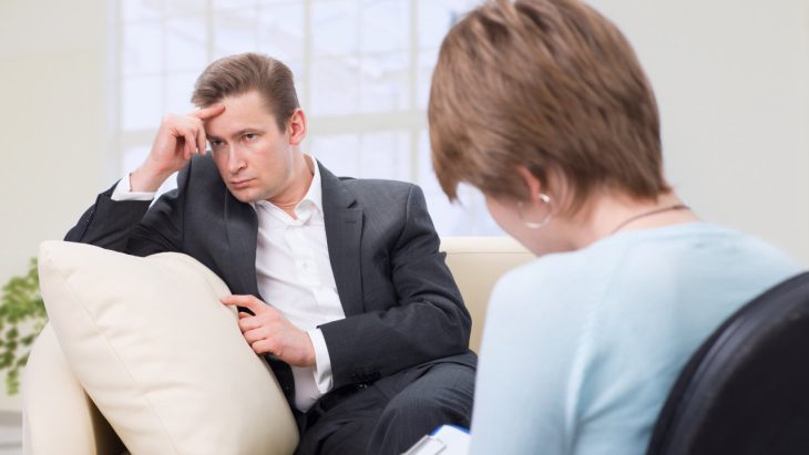 A stressed looking man sits on a couch, speaking with a doctor about relapse prevention.