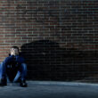 A man sitting against a brick wall, casting a long shadow