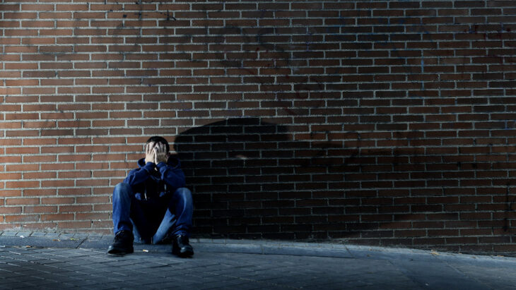 A man sitting against a brick wall, casting a long shadow