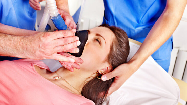 A woman lies in a hospital bed while a doctor places a breathing mask on her face to administer anesthesia, part of a rapid opiate detox regimen