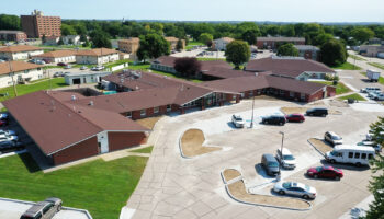 Exterior aerial view showing brick building with brown roof, parking lot with vehicles, and surrounding landscaping