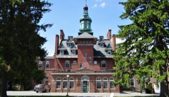 Exterior of brick Victorian-style building with green cupola tower, mansard roof, and manicured grounds with mature trees