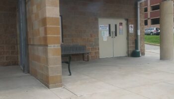 Exterior entrance showing brick building facade, covered portico with metal beams, cream-colored double doors with facility signage, and concrete walkway