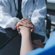 A doctor holds the hand of a patient, comforting them. Learn more about the signs and health risks of alcoholism in women here.
