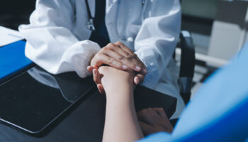 A doctor holds the hand of a patient, comforting them. Learn more about the signs and health risks of alcoholism in women here.