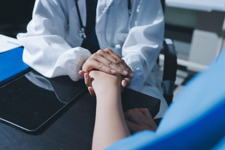A doctor holds the hand of a patient, comforting them. Learn more about the signs and health risks of alcoholism in women here.