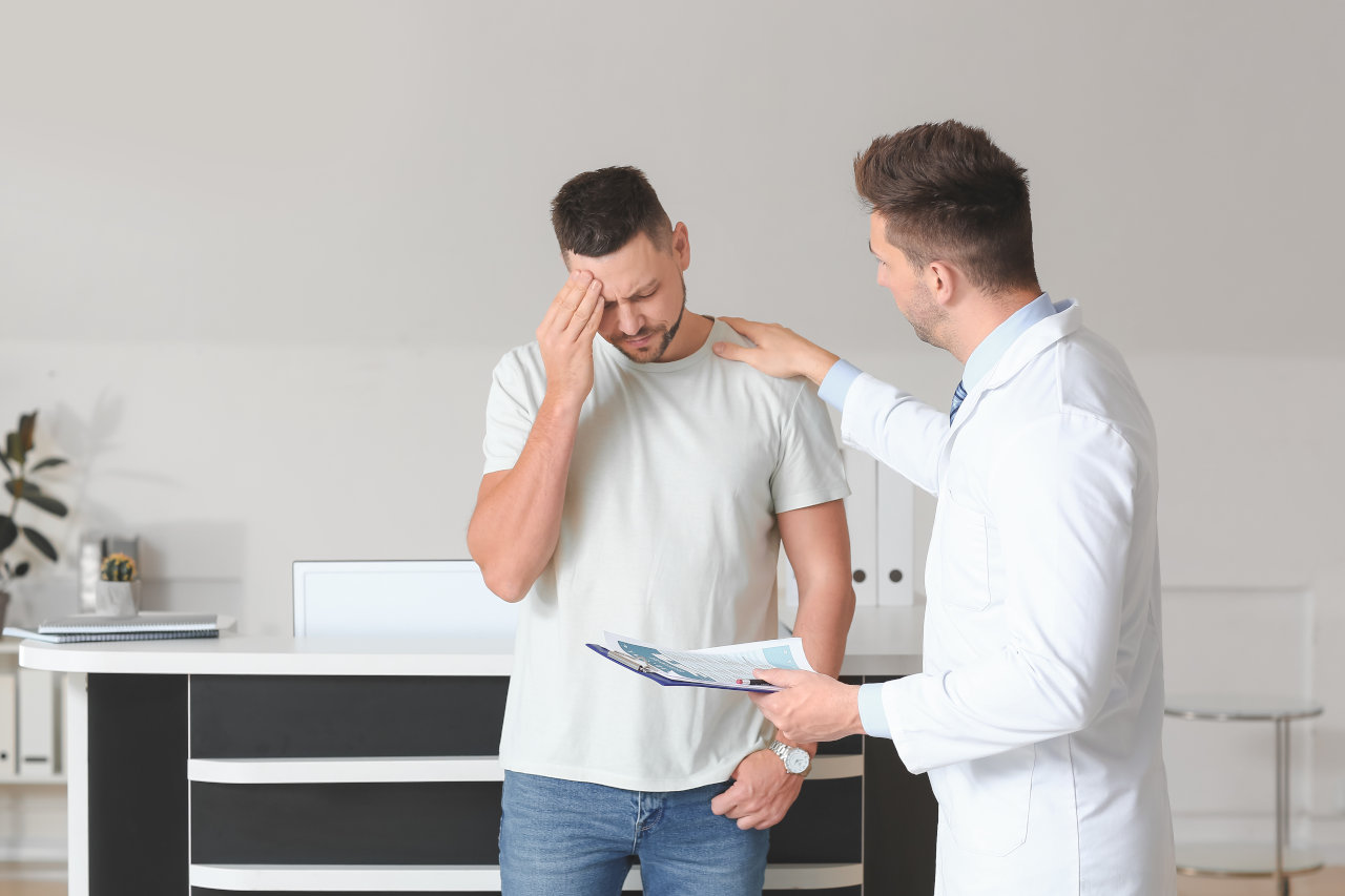 A man grips his head in pain while speaking with a doctor. Learn more about Xylazine (tranq) side effects, overdose risks, and detox help here