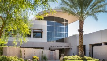 Exterior view of modern facility building with large glass windows, stone accent wall, landscaped grounds, and palm trees