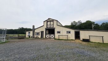 Exterior cream and dark-trim barn structure with large doors, metal fencing, gravel courtyard and pastoral setting