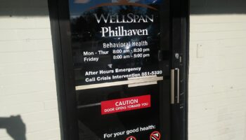 Exterior entrance door with WellSpan Philhaven Behavioral Health signage displaying hours of operation and emergency contact information