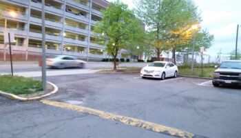 Exterior view of multi-story brick facility with parking area, vehicles, and mature trees