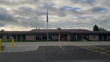 Exterior front view of medical facility building with accessible parking spaces marked in yellow, flagpole, and cloudy sky