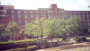Exterior view of large red brick hospital building with multiple stories, windows, and surrounding landscaped grounds with trees and pathways