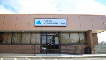 Exterior front entrance of Ocean Monmouth Care showing illuminated signage, brick facade, glass doors, and accessible ramp