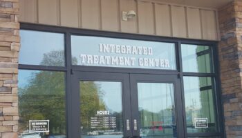 Exterior view of treatment center main entrance with glass doors, signage reading 'Integrated Treatment Center', and operating hours posted on window