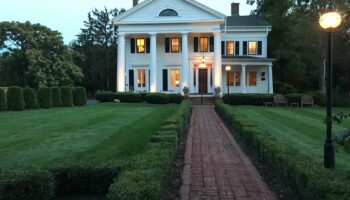 Exterior view of white colonial-style building with columns, manicured landscaping, brick pathway, and evening lighting