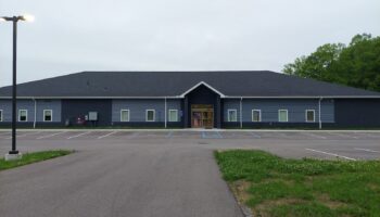 Exterior view of single-story treatment facility with dark blue and gray siding, central entrance, accessible parking spaces, and landscaped grounds