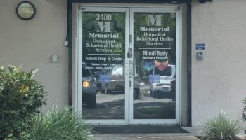 Exterior entrance showing glass double doors with signage for Memorial Outpatient Behavioral Health Services, Rebels Drop-In Center, and Mind/Body Wellness Program