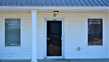 Exterior front entrance of Outlook Recovery treatment center showing white building facade with signage, central entrance door, and unit number 10