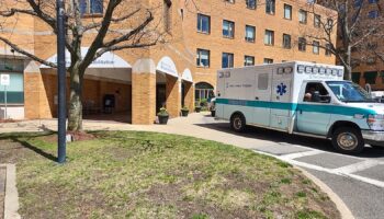 Exterior view of brick building entrance with arched awning, ambulance parked in bay, and landscaped courtyard with trees