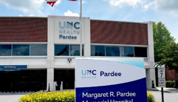 Exterior front entrance showing UNC Health Pardee building facade with blue signage, parking garage, and American flag