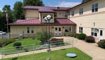 Exterior view of treatment facility showing cream-colored building with burgundy metal roof, landscaped grounds, patio area, and American flag