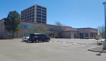 Exterior view of La Clinica de Familia medical facility with tan building facade, parking area, and multi-story structure
