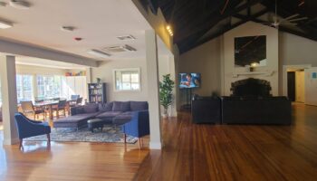 Interior common space with hardwood floors, dark seating, fireplace, television, and large windows with natural light