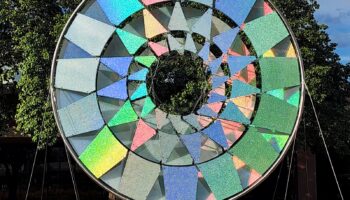 Exterior view of colorful geometric wind sculpture with rotating panels in multiple colors against blue sky and surrounding vegetation