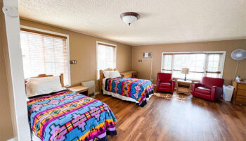 Interior patient bedroom with two beds featuring colorful patterned comforters, wooden flooring, and multiple windows with natural light