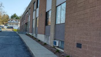 Exterior side view of brick facility building with large windows, concrete walkway, and surrounding landscaping and parking area