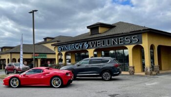 Exterior front facade of Synergy Wellness showing main entrance with yellow building, signage, and visitor parking with landscaping