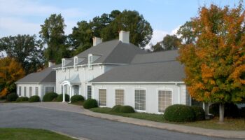 Exterior view of white colonial-style building with gray roof, columns, multiple windows, and landscaped grounds with trees