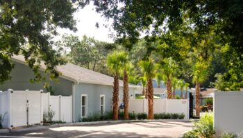 Exterior gated entrance showing white facility walls, palm trees, and paved driveway with landscaping