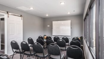 Interior meeting room with rows of black chairs, whiteboard, barn door, and large windows providing natural light.