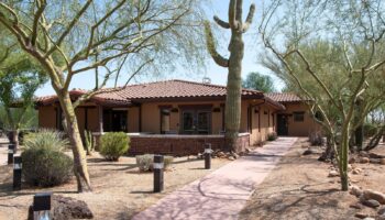 Exterior of main facility building with tan stucco walls, tiled roof, surrounded by native desert plants and saguaro cacti