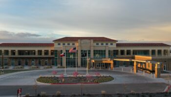 Exterior of VA Lubbock Clinic showing tan multi-story building with columned entrance, red tile roof, American flag, and landscaped roundabout at dusk