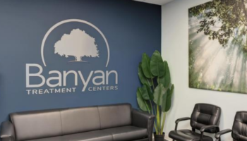 Interior facility lobby with dark blue accent wall displaying large Banyan Treatment Centers tree logo, gray sofa, chairs, and potted plant