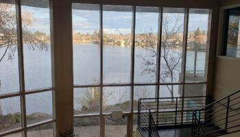 Interior space with expansive windows overlooking calm water, landscaping, and far shoreline with trees and buildings visible.